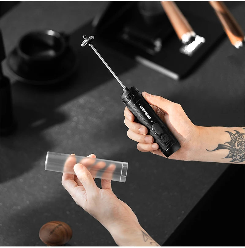 Handheld black electric milk frother with a slim metallic whisk, shown with a clear protective sleeve.