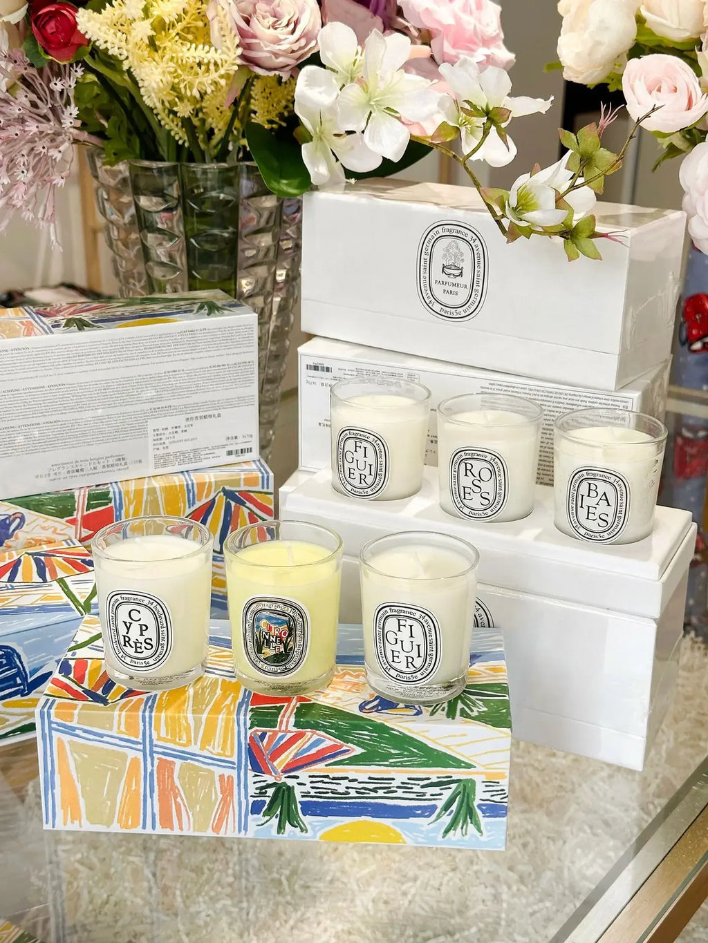 A collection of small white scented candles in glass jars, displayed with colourful decorative gift boxes and flowers.