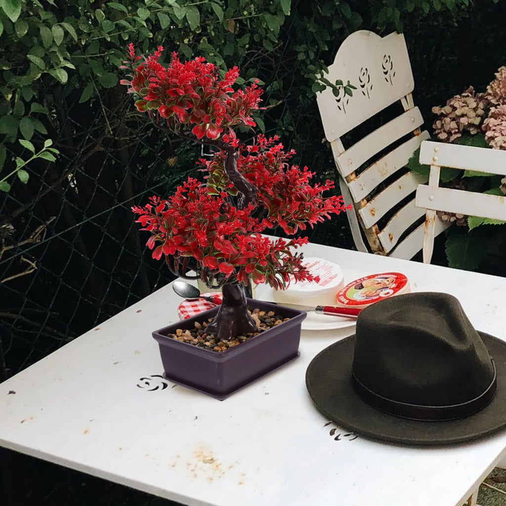 Artificial bonsai tree with bright red leaves in a rectangular brown pot on a white outdoor table with a fedora.