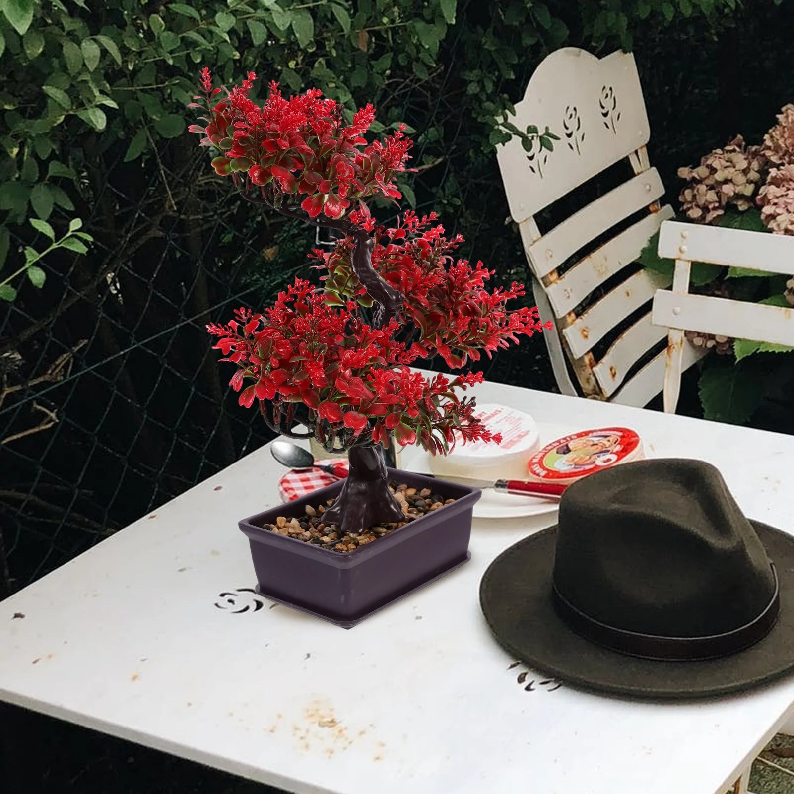 Artificial bonsai tree with bright red leaves in a rectangular brown pot on a white outdoor table with a fedora.