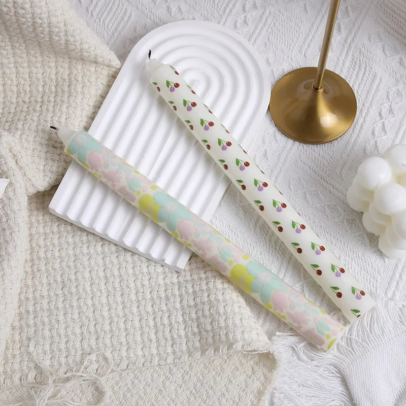 Two tapered candles on a white tray; one with a pastel floral pattern, the other with a red and purple cherry print.