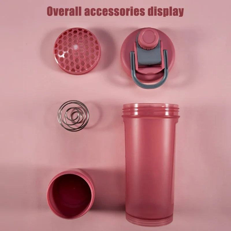 Disassembled pink protein shaker bottle with a honeycomb filter, metal whisk ball, and locking lid on a pink background.