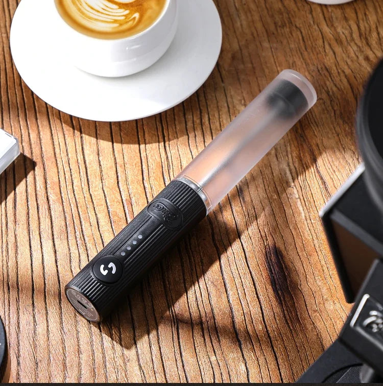 Portable black electric milk frother with a translucent wand, battery indicators, and power button on a wooden table.