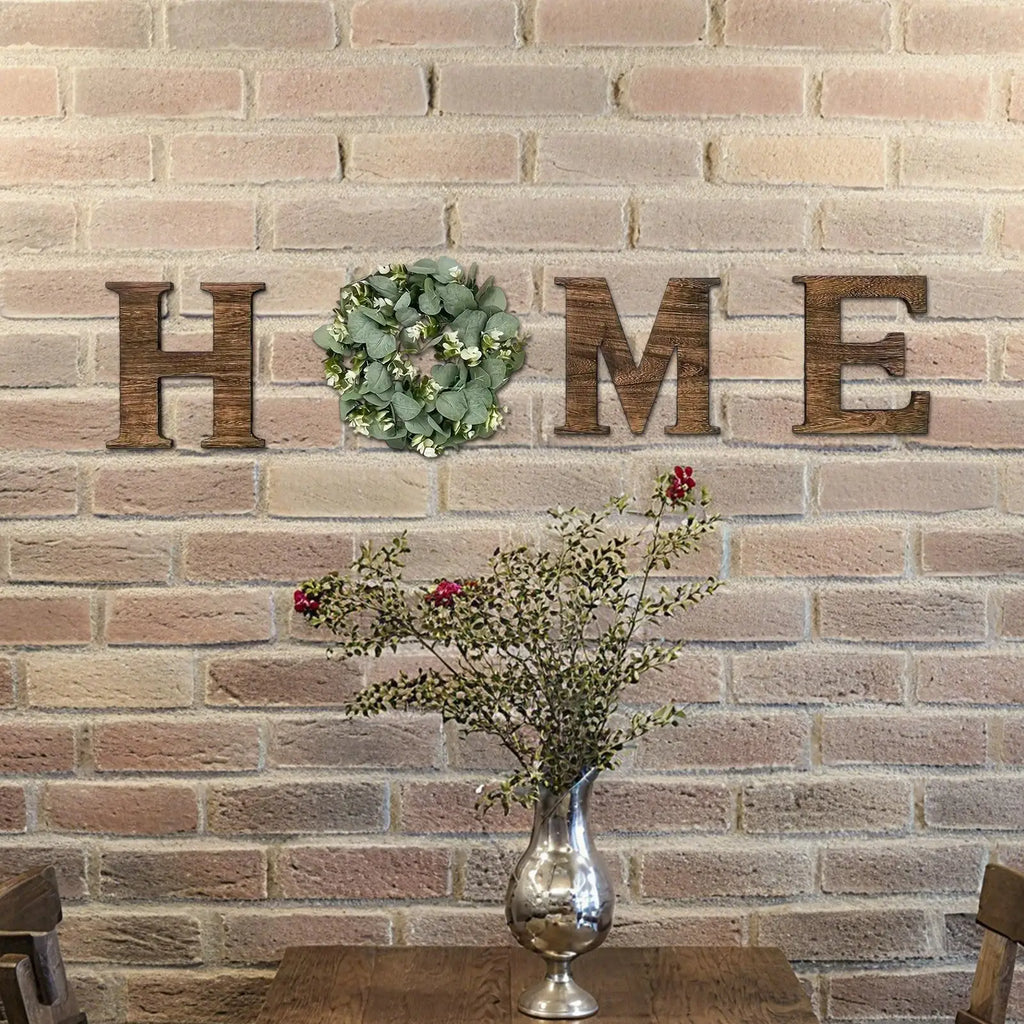Rustic wooden 'HOME' wall sign with an artificial eucalyptus wreath replacing the 'O', mounted on a brick wall.