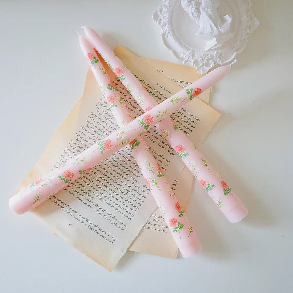 Three light pink taper candles decorated with delicate floral rose patterns, resting on vintage book pages.
