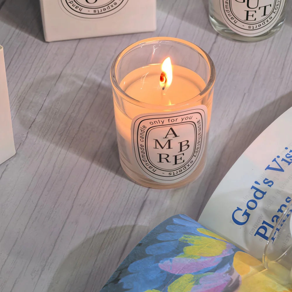 Lit scented candle in a clear glass jar with a white oval label, resting on a light grey wood-effect surface.
