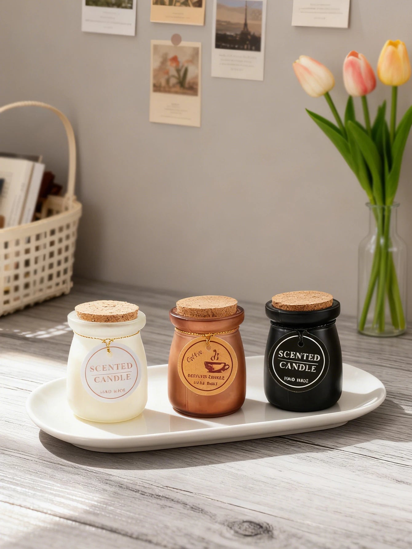 Three small scented candles in white, bronze, and black glass jars with cork lids, displayed on an oval ceramic tray.