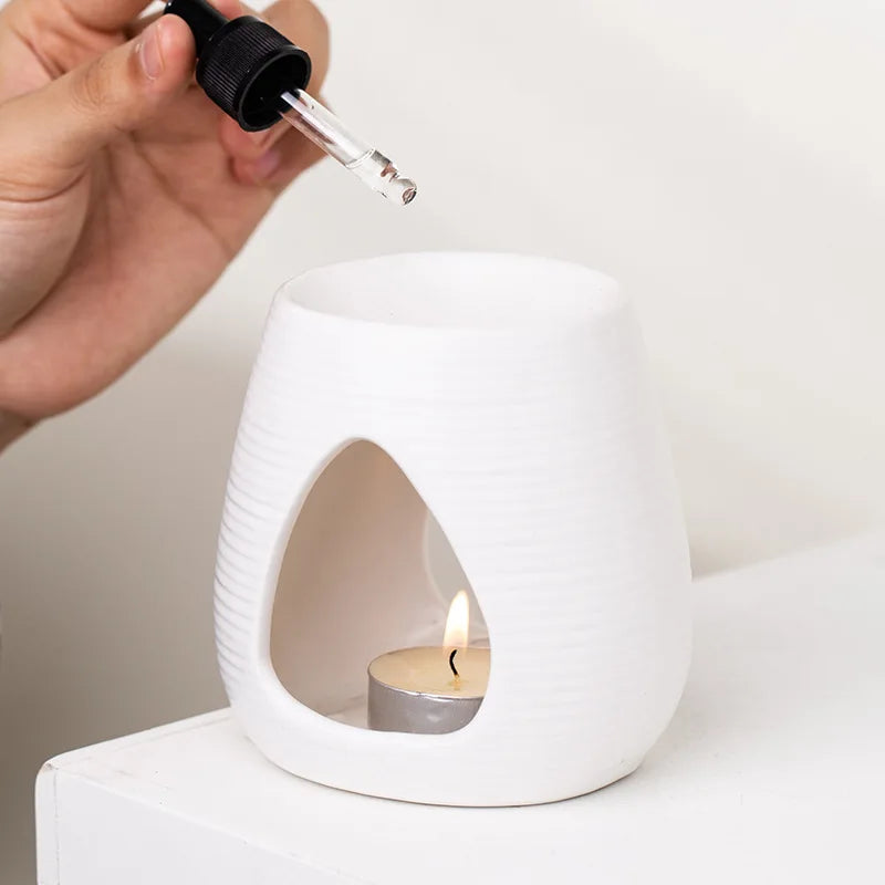 White ceramic oil burner with ribbed texture and teardrop cutout, featuring a lit tealight and a hand holding a dropper.