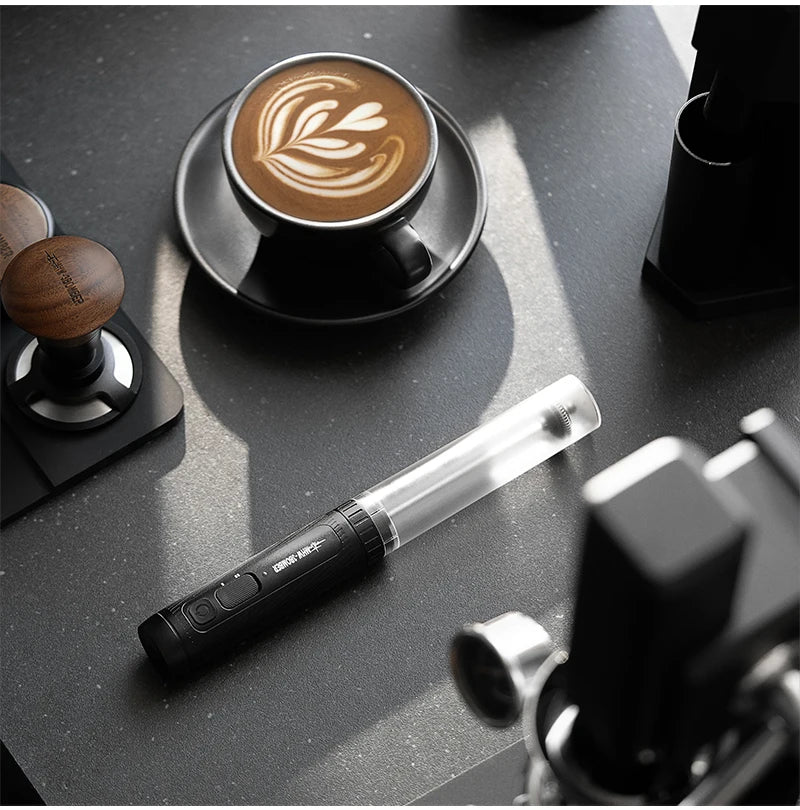 Black handheld electric milk frother with a clear plastic housing, lying on a dark, modern coffee station surface.