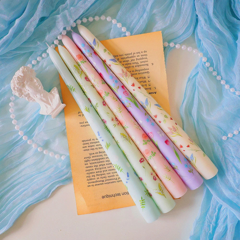Five pastel-coloured tapered candles featuring delicate floral prints, arranged on paper with blue fabric and pearls.