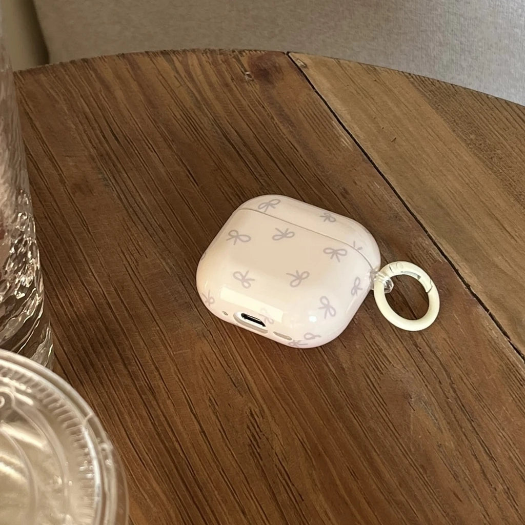 Cream AirPods Pro case with a grey bow pattern, smooth plastic finish, and integrated keyring on a wooden table.