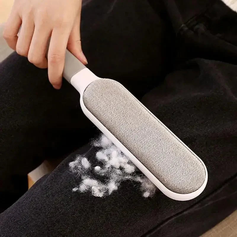 A hand using a grey and white lint remover brush on black trousers to pick up white lint.