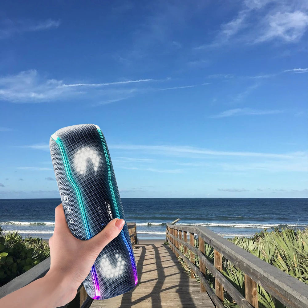 Handheld, cylindrical grey portable speaker with vibrant LED light accents against a coastal beach background.