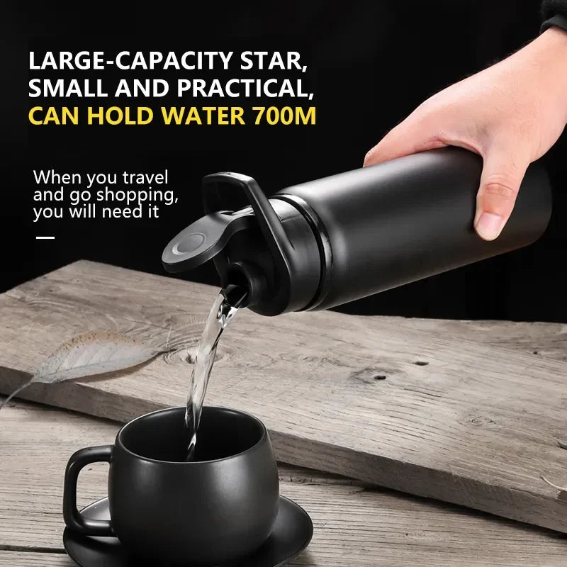 A hand pouring water from a matte black 700ml travel bottle into a matching black cup on a rustic wooden table.