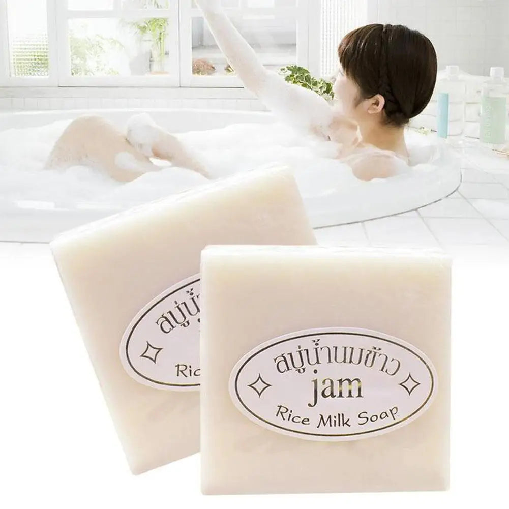 Two square bars of off-white rice milk soap with oval labels, set against a blurred background of a woman in a bath.