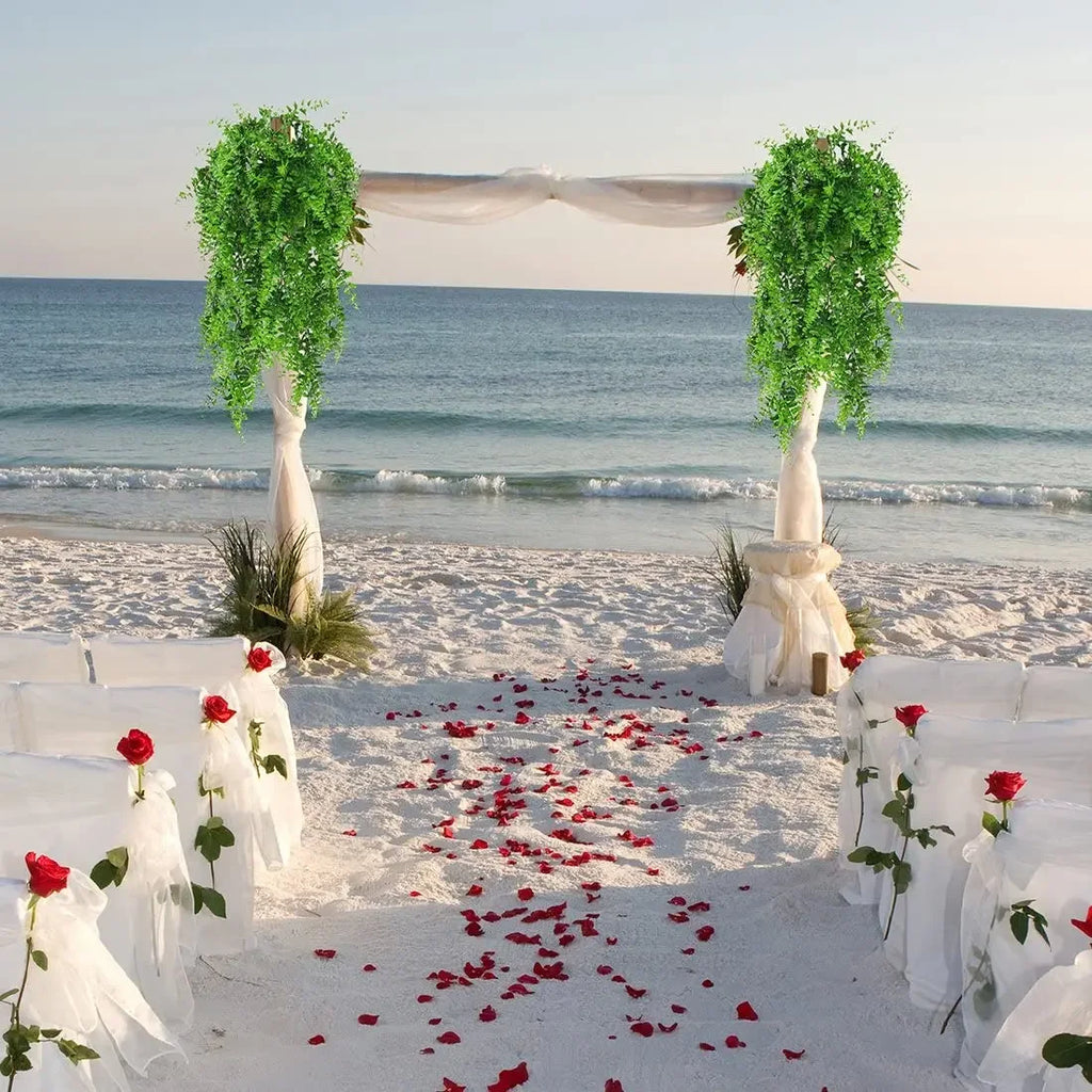 Two lush green artificial Boston fern bushes hang from a wedding arch on a sandy beach.