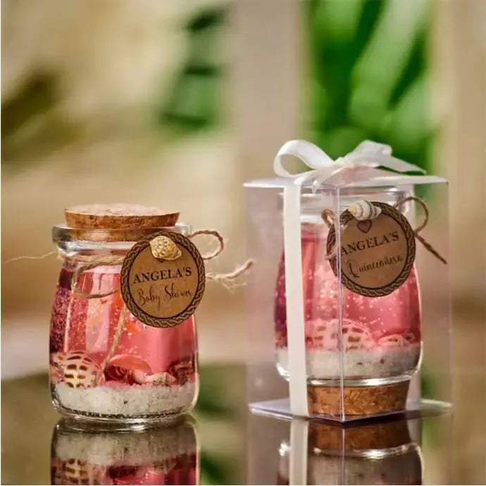 Glass jars with cork lids, pink liquid, sand, and seashells, featuring rustic tags and a clear gift box with ribbon.