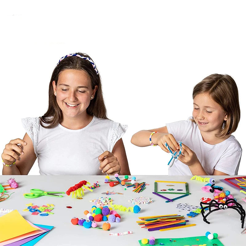 Two girls creating colourful crafts with pom-poms, pipe cleaners, beads, and craft sticks on a white table.