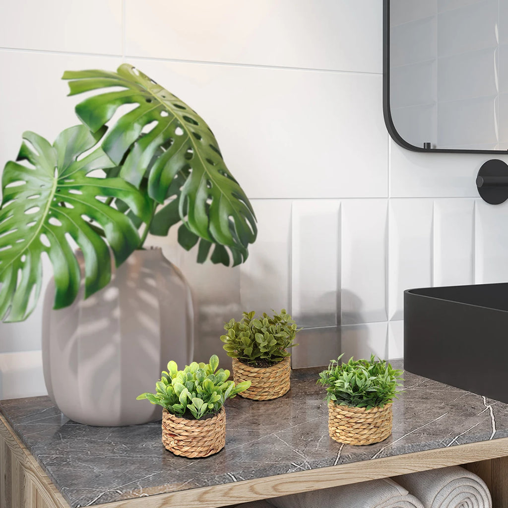 Three small artificial plants in woven rattan pots sit on a marble vanity next to a large monstera plant.