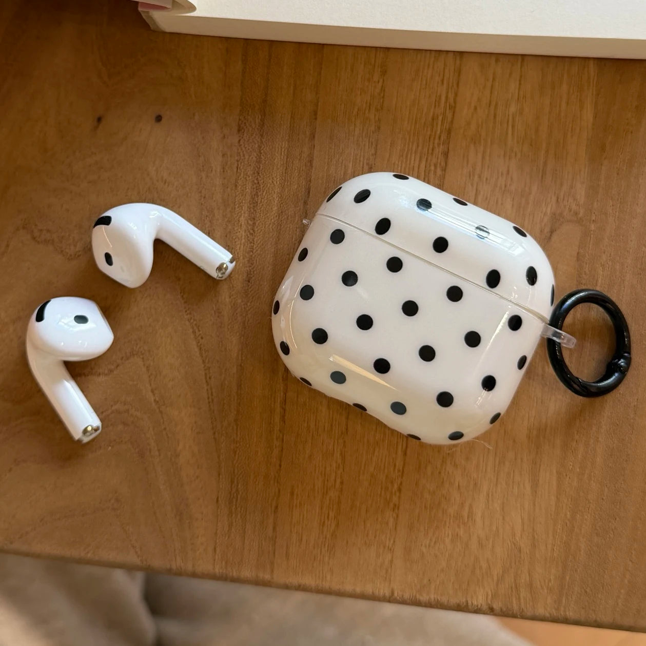 White polka dot protective hard case for AirPods, featuring a black carabiner clip on a wooden table.
