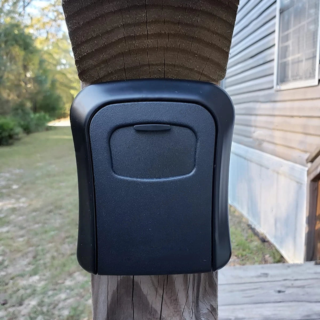 A matte black, rectangular key lock box with a protective cover, mounted on a wooden outdoor post.
