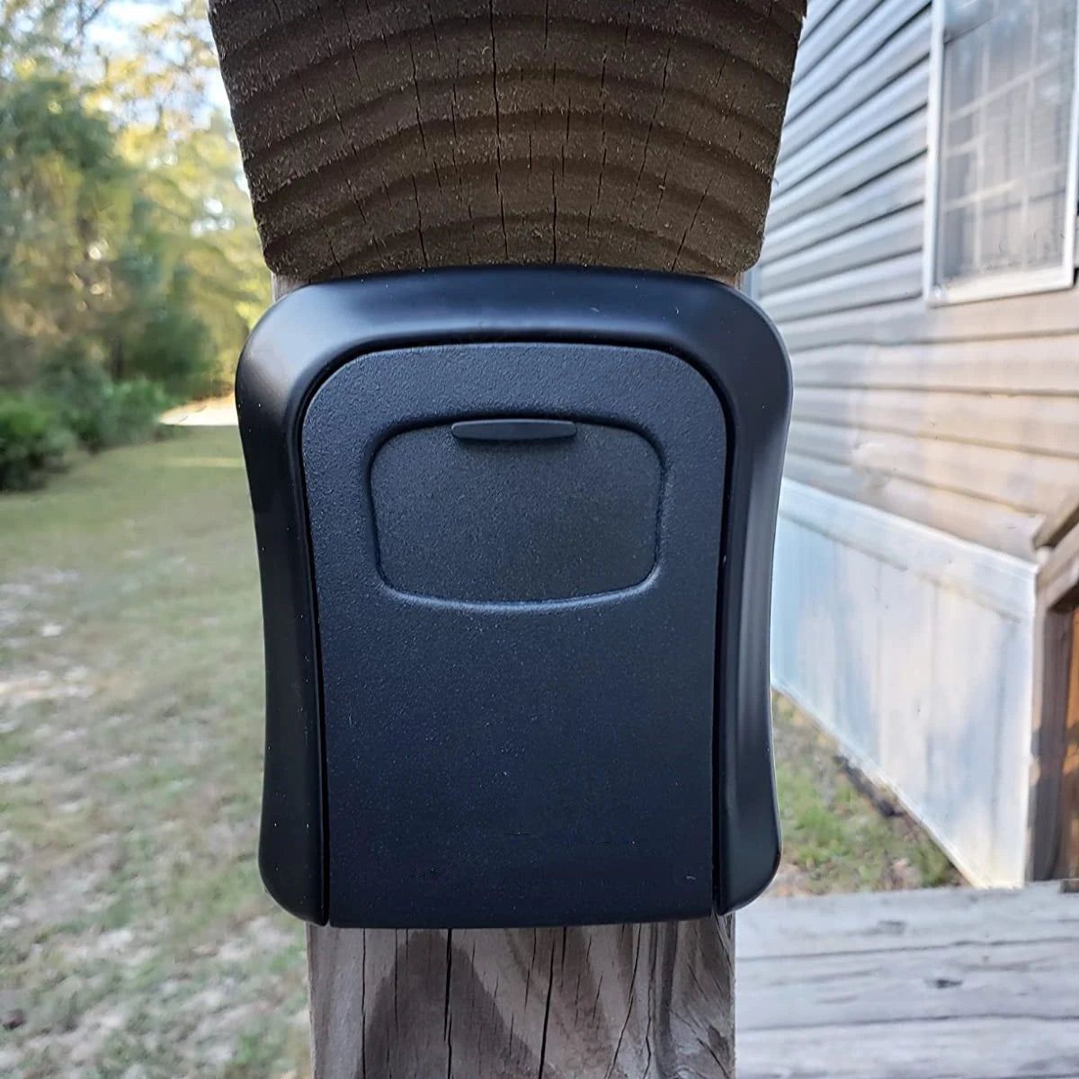 A matte black, rectangular key lock box with a protective cover, mounted on a wooden outdoor post.