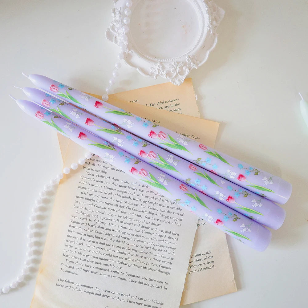 Three tapered lilac dinner candles decorated with delicate pink tulips and small blue flowers.