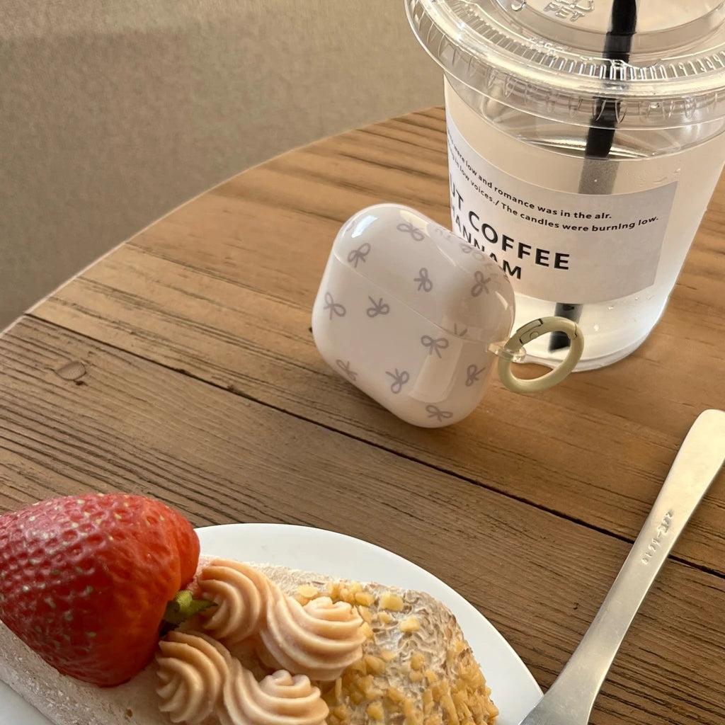 Glossy white protective AirPods case with a grey bow print and integrated side ring on a wooden cafe table.