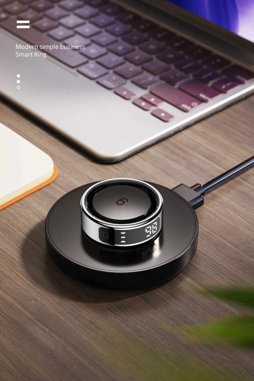 Sleek silver and black smart ring with a digital display, resting on a wireless charging pad on a wooden desk.