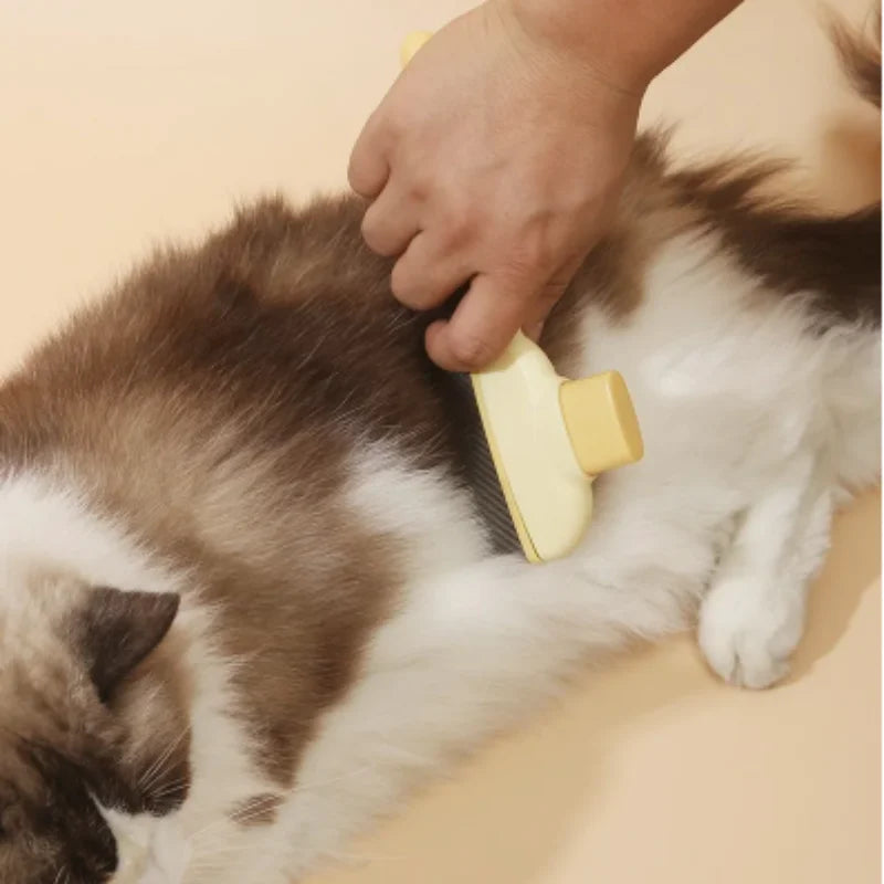 A person uses a yellow self-cleaning slicker brush to groom a fluffy brown and white cat on a cream background.