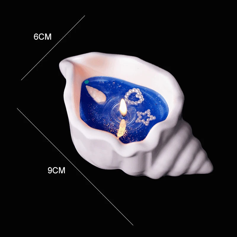 White seashell-shaped candle with blue wax, decorative pearls, and a lit wick. Dimensions: 6cm by 9cm.