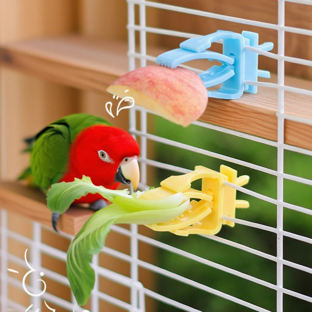 A red-headed green parrot eating fruit and vegetables held by blue and yellow plastic bird cage food clips.