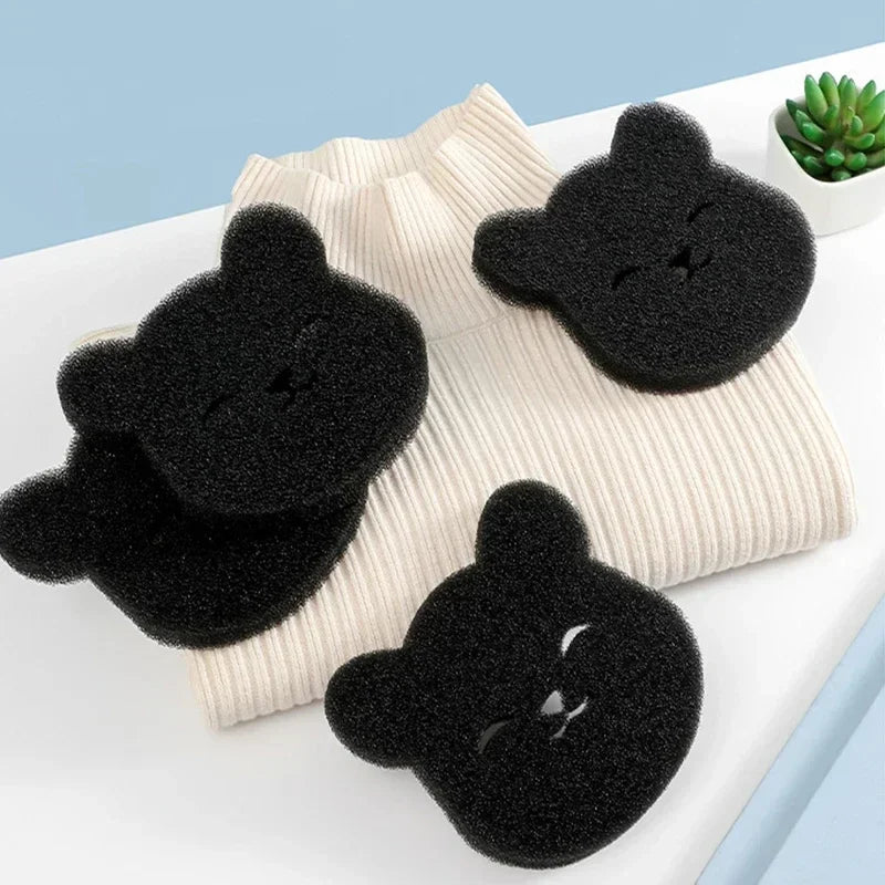 Three black, bear-shaped scrubbing sponges with cute smiling faces resting on a folded cream-coloured ribbed jumper.