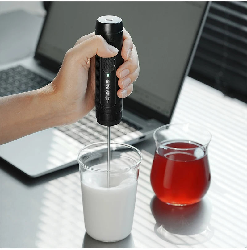 Handheld black electric milk frother with a sleek, minimalist design mixing milk in a glass cup.