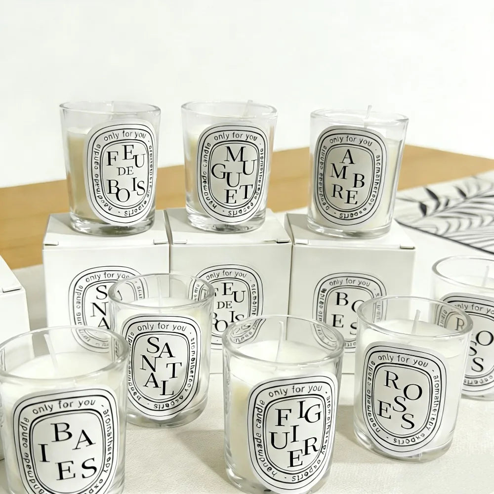 A collection of white wax candles in clear glass tumblers, each with a distinctive oval label and individual scent name.