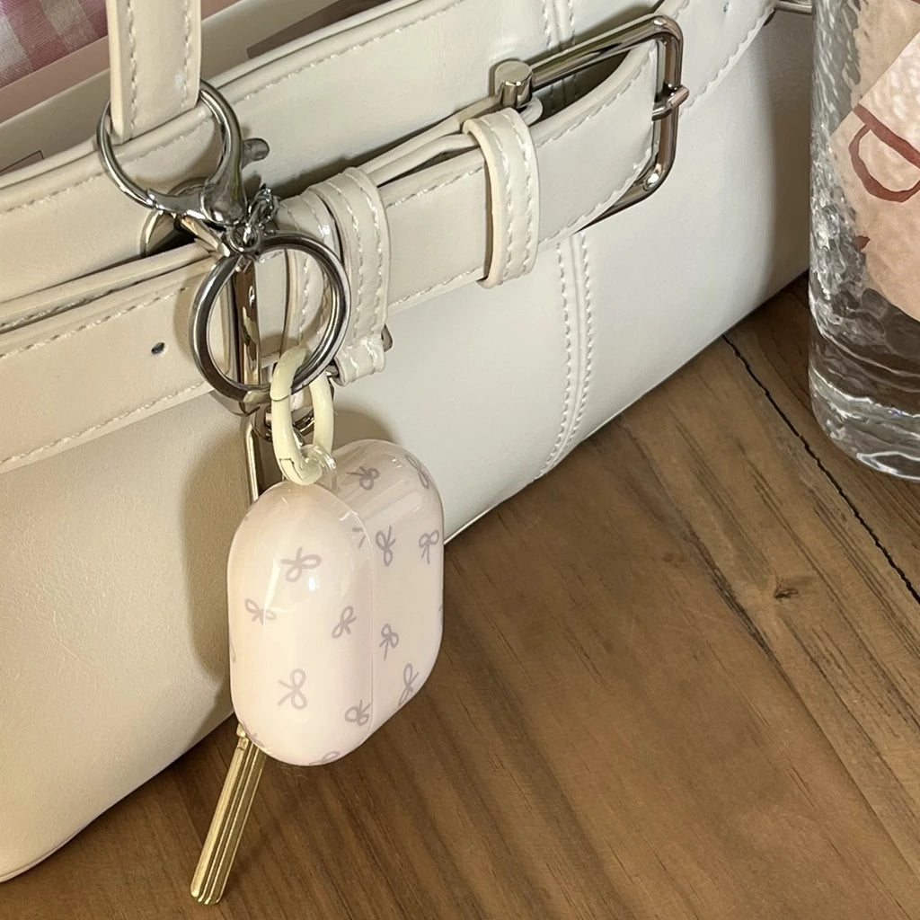 Pale pink, bow-print protective AirPods case in smooth silicone with a silver metal keyring clip.