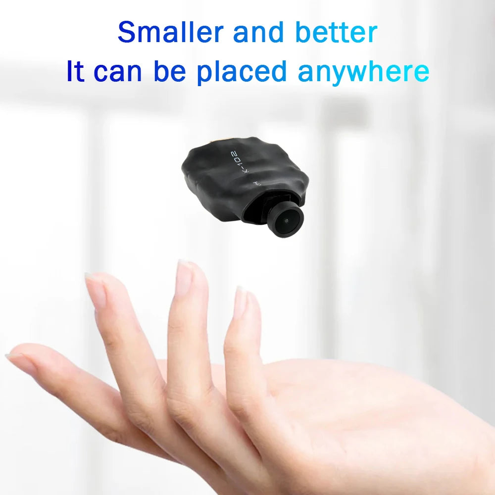 Small black K-102 micro security camera with a protruding lens, held above a hand.