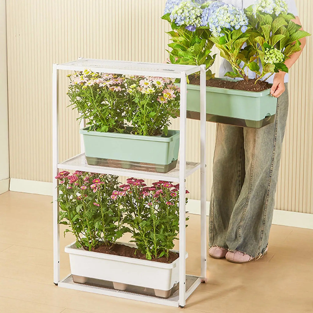 A white metal plant stand holds rectangular, sage green and white self-watering planters filled with blooming flowers.