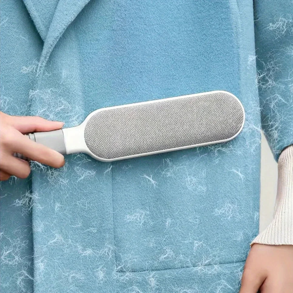 A hand uses a grey and white lint remover brush on a light blue wool coat covered in loose fibres.
