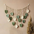 Boho macrame wall hanging featuring sage green and cream leaf-shaped pendants suspended on a beaded string.