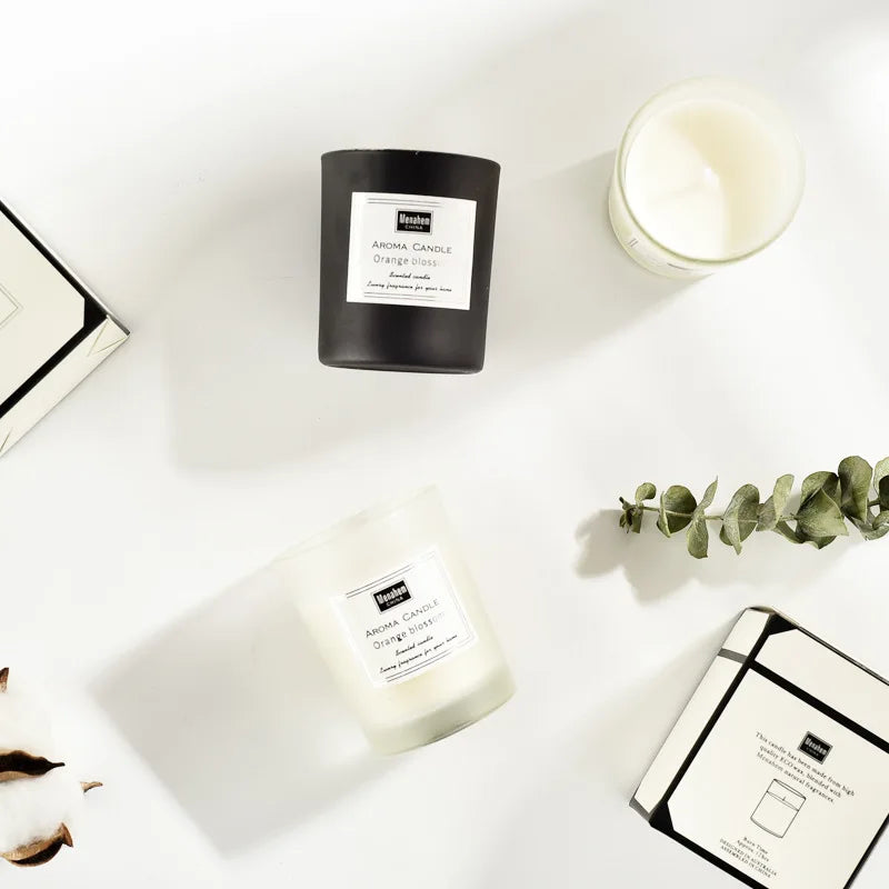 Two glass aroma candles, one matte black and one clear, presented on a white surface with decorative greenery.