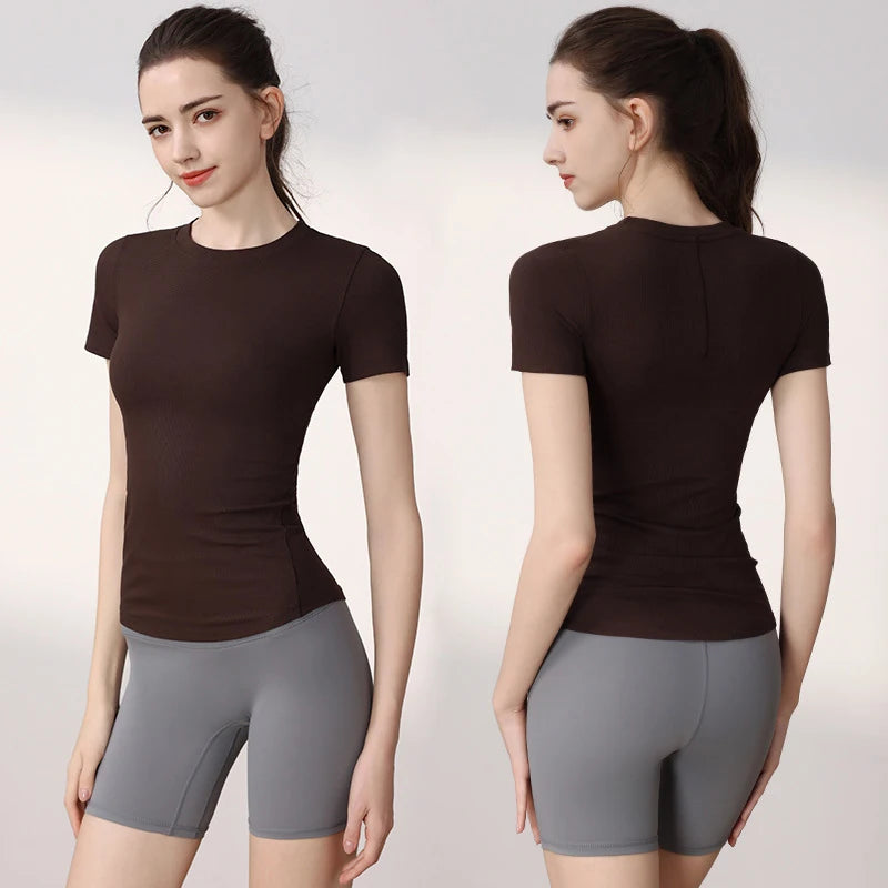 A model wears a fitted, dark brown ribbed short-sleeve sports top paired with grey athletic shorts.