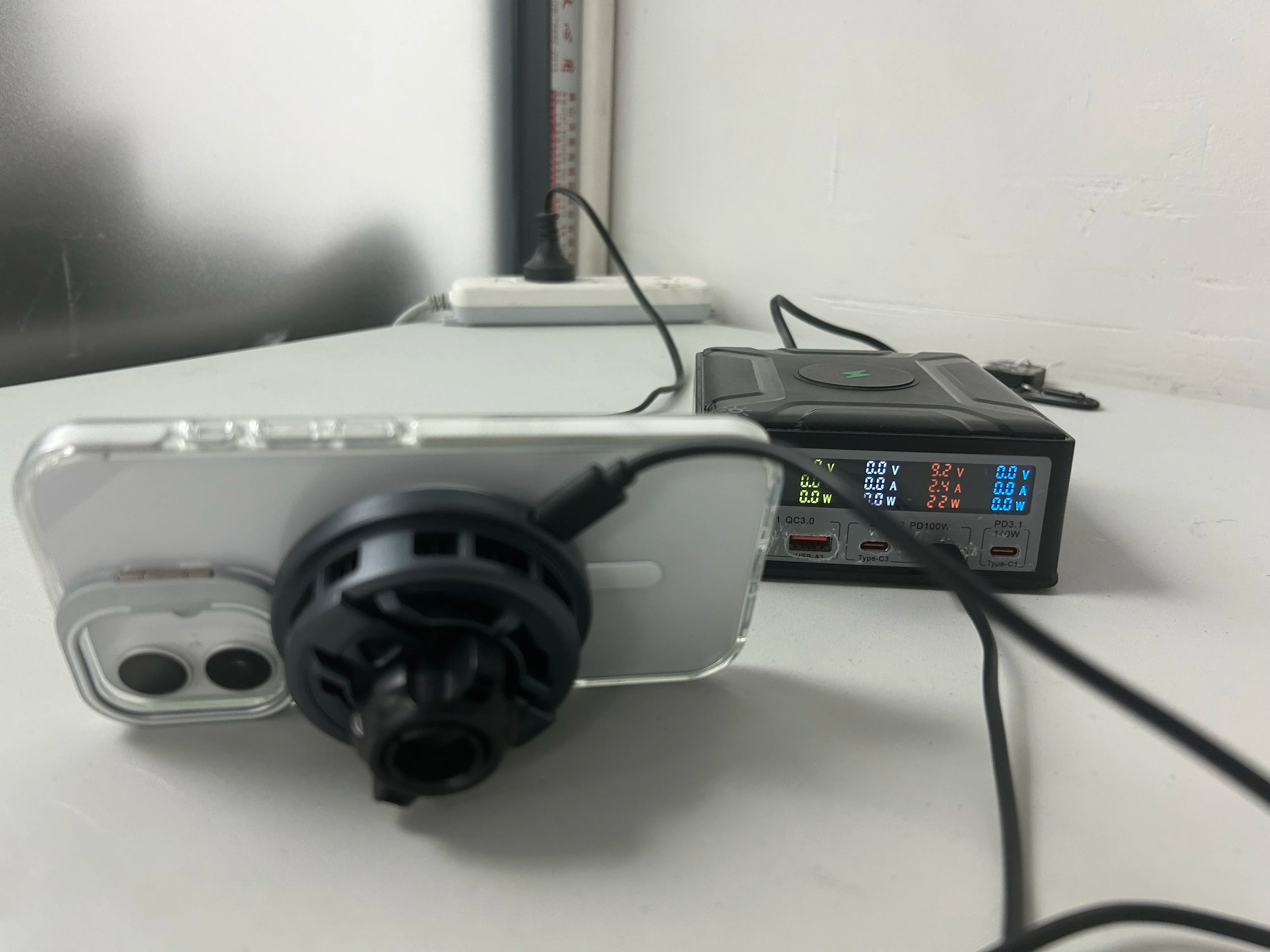 Smartphone with magnetic cooling fan attached, connected to a black digital display charging station on a white desk.