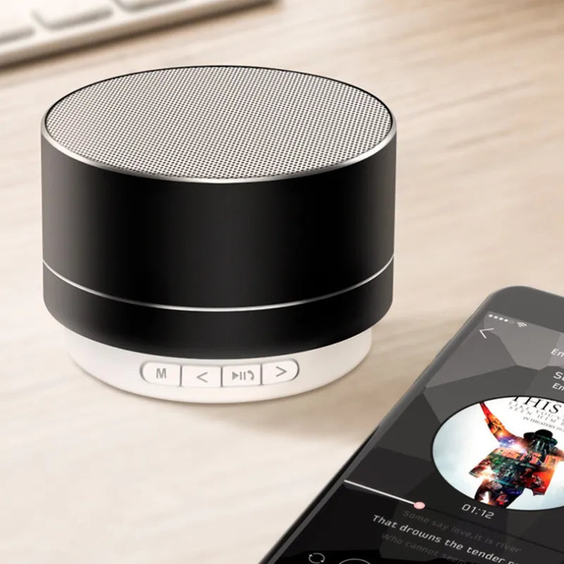 Portable black and silver cylindrical Bluetooth speaker with playback buttons, sitting next to a smartphone.