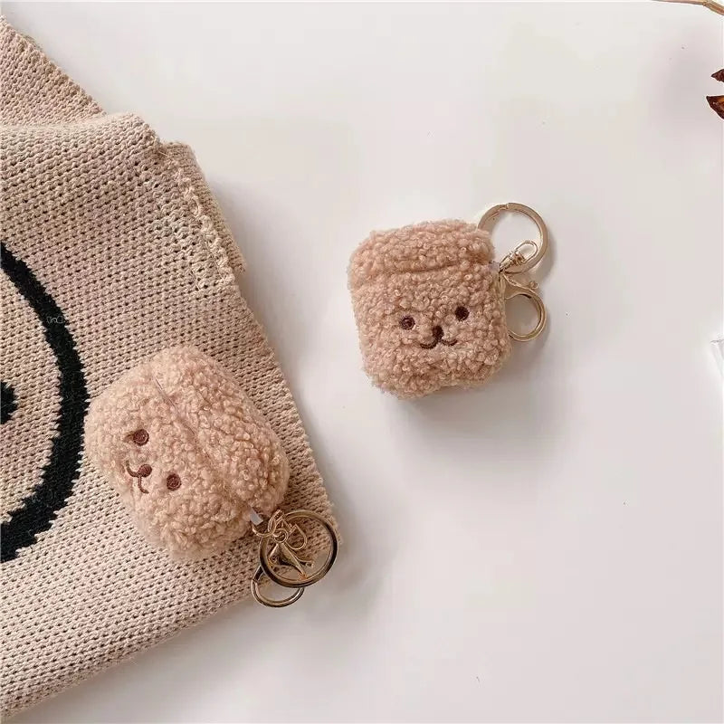 Beige teddy-fleece AirPods case with an embroidered bear face and gold-tone keychain ring.