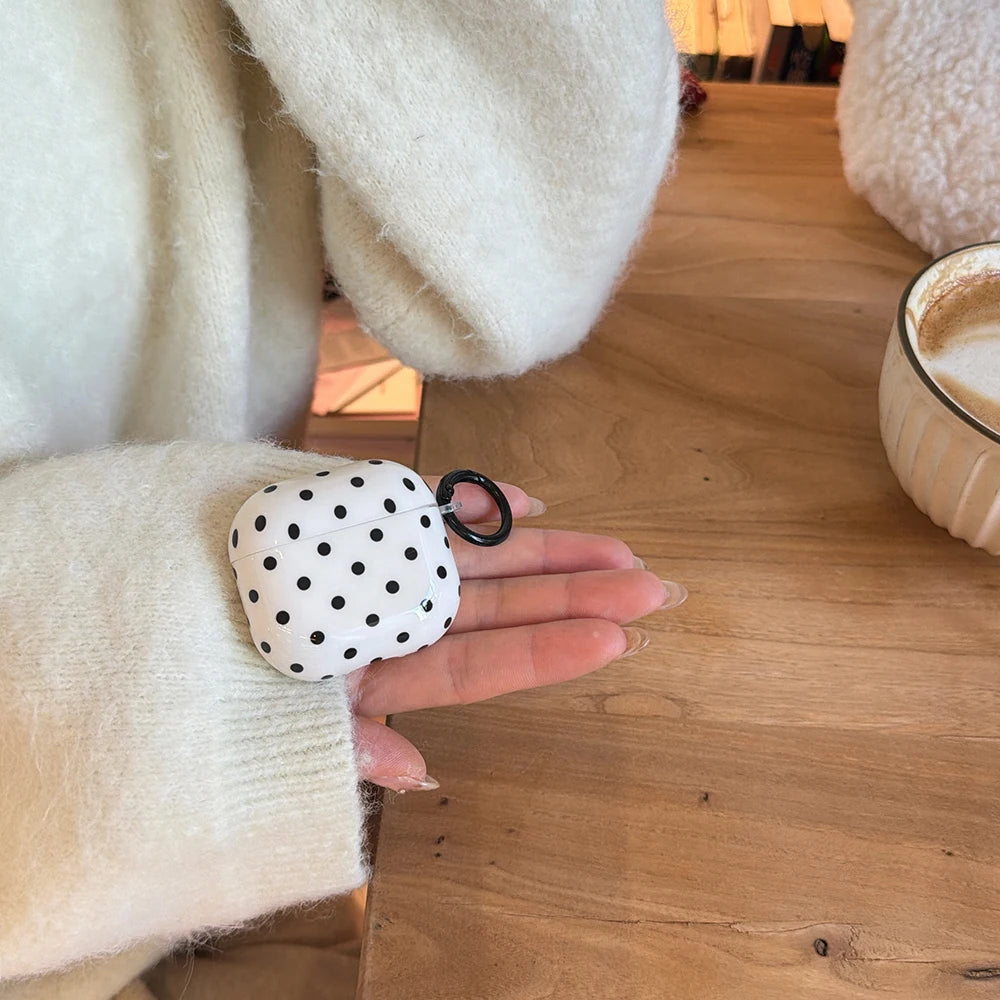 White polka dot AirPods case with black keyring attachment, held in a hand. Glossy plastic finish.