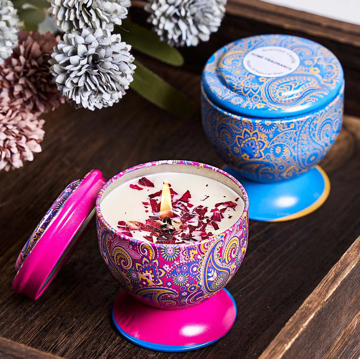 Burning scented candle in a decorative, patterned pink tin with a matching base, next to a blue paisley tin.