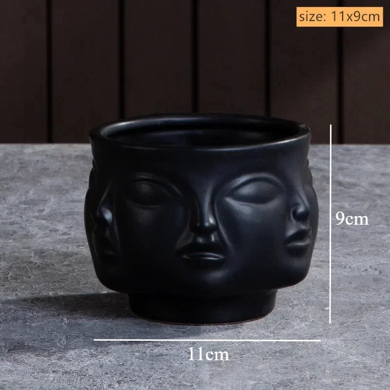 Matte black ceramic plant pot featuring a sculpted, multi-face abstract design. Measures 11cm diameter by 9cm height.