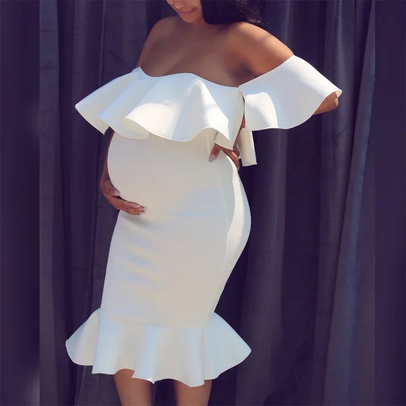 White off-shoulder maternity dress with a ruffled neckline and hem, form-fitting silhouette.