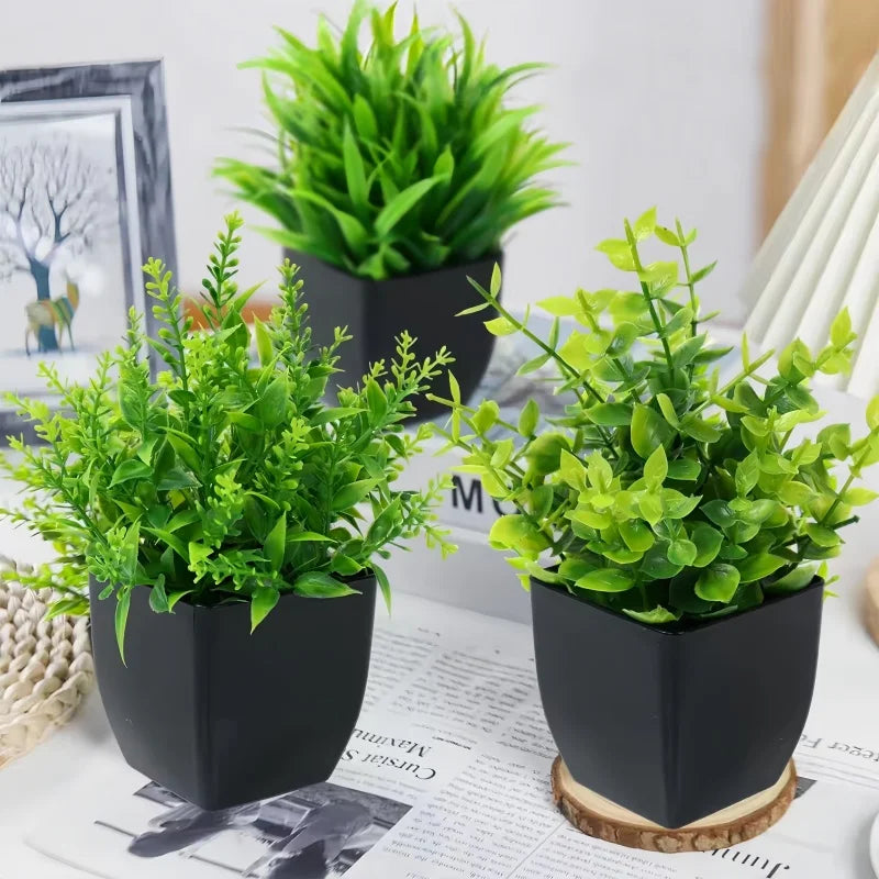 Three artificial green plants in modern, matte black square pots arranged on a table with decor.